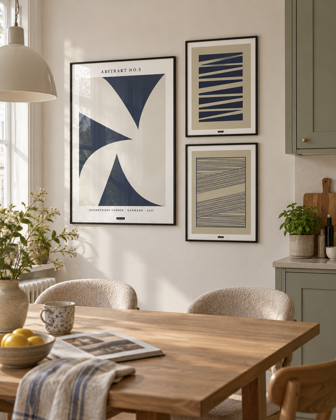 Blå former billedvæg med tre geometriske plakater i sorte aluminiumsrammer i et dansk køkken-alrum med sagegrønne skabe.