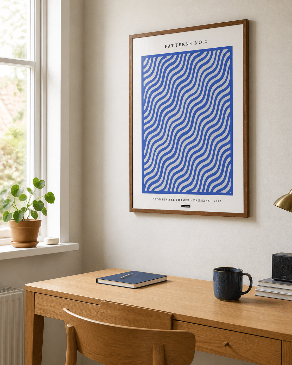 Patterns No.2-plakaten med blå bølger i en varm træramme hænger på en lys væg i et naturligt dansk hjemmekontor med skrivebord og dagslys.